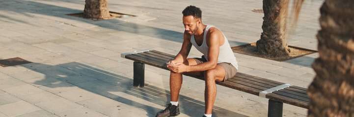 Obraz premium Young bearded male athlete is resting after training, sitting on bench, using smartphone, panorama