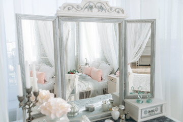 Elegant vanity setup featuring ornate mirror reflecting white canopy bed with pastel pillows. Decorated with flowers, candelabra, and delicate items, soft natural light adds a peaceful ambiance