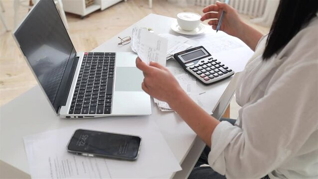 Side view of woman working with financial papers on white office desk with laptop, counting on calculator, paying bills, planning budget to save some money. Independent accounting, remote accountant