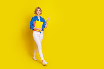Full body portrait of pretty young lady walk book v-sign empty space wear shirt isolated on yellow color background