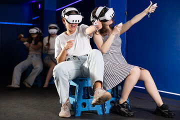 Excited young female friends having fun in modern virtual reality room, playing pair versus pair game in VR glasses, manipulating objects or shooting at targets with gaming controllers in hands..
