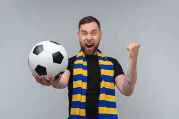 Studio shot of euphoric soccer supporter man wearing yellow-blue scarf posing with a ball in hand over grey background making winners gesture clenching his fist and reaching the ball to the camera