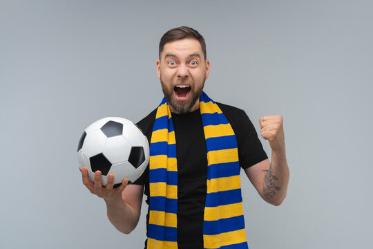 Studio portrait of euphoric soccer supporter man wearing yellow-blue scarf posing with a ball in hand over grey background making winners gesture clenching his fist