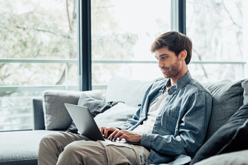 Handsome serious man sits on sofa with computer on laps, spend time at home web surfing information, use modern tech, make order on internet. Freelancing, telework, new software learning, tech concept