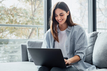 Concentrated millennial girl sit on couch working on laptop browsing internet at home during lazy weekend, focused young woman freelancer busy using computer surfing wireless web shopping online.