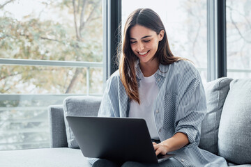 Concentrated millennial girl sit on couch working on laptop browsing internet at home during lazy...