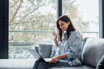 Smiling girl sit on couch at home wave talk with friend or family having video call on laptop, happy young woman greeting say hello, speak communicate using dating online application on computer.