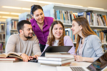 Fototapeta premium Group of positive friendly international university students working on joint research in library, looking for information in books and websites using laptop