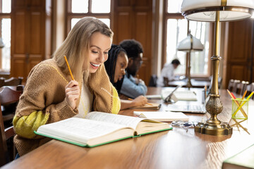 Blonde young woman in beige studying in the library and looking excited