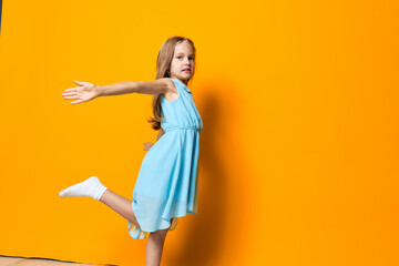 Joyful girl in blue dress balances with arms outstretched on a bright orange wall under a clear sky