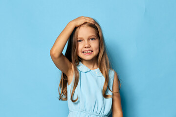 Little girl in a blue dress standing with hands on her head against a matching blue background