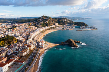 Scenic drone view of coastal Catalan town of Blanes overlooking white residential buildings and Sa Palomera Rock on background of San Juan hill with ancient castle on sunny day, Barcelona, Spain ..