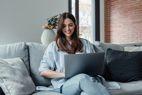 Concentrated millennial girl sit on couch working on laptop browsing internet at home during lazy weekend, focused young woman freelancer busy using computer surfing wireless web shopping online.