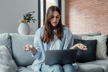 Angry young woman sit on sofa in living room frustrated having operational problems working on...