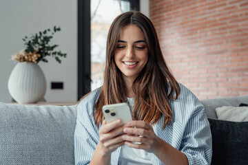 Smiling woman looking at smartphone screen, sitting on couch, enjoying leisure time with mobile...