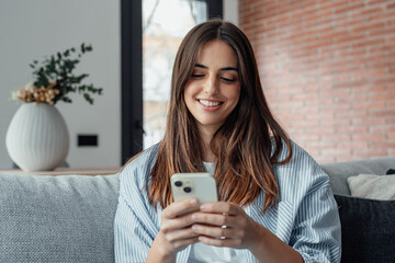 Smiling woman looking at smartphone screen, sitting on couch, enjoying leisure time with mobile...