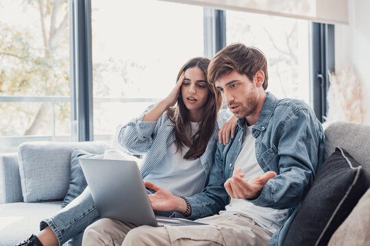 Millennial couple annoyed by stuck laptop or online news sitting together on sofa, frustrated young angry customers confused by computer problem feeling perplexed or puzzled about email message.