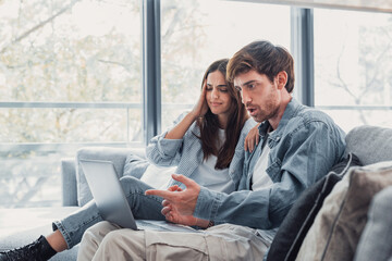 Millennial couple annoyed by stuck laptop or online news sitting together on sofa, frustrated young...