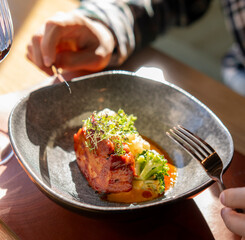 A close-up photo showcasing a delicious gourmet meal on a restaurant table, with hands holding utensils and a glass of wine nearby