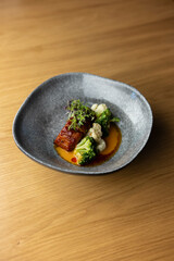 A grey bowl filled with a delicious dish of roasted pork belly with cauliflower, broccoli, and a tangy orange sauce, placed on a wooden table