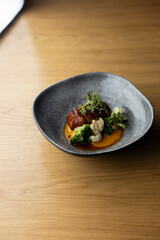 A grey bowl filled with a delicious dish of roasted pork belly with cauliflower, broccoli, and a tangy orange sauce, placed on a wooden table
