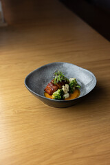 A grey bowl filled with a delicious dish of roasted pork belly with cauliflower, broccoli, and a tangy orange sauce, placed on a wooden table