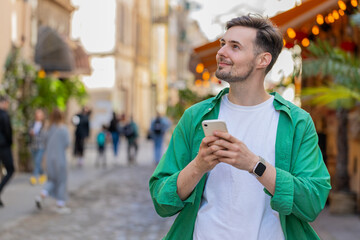 Caucasian young man using smartphone typing texting social media messages e-mail looking for a way on map in mobile navigator app outdoors. Smiling traveler walking on city street. Town lifestyles.