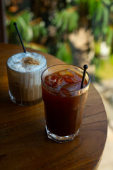 Iced americano and buttercloud latte coffee served together on transparent glass, over wooden table surface, in cafe environment