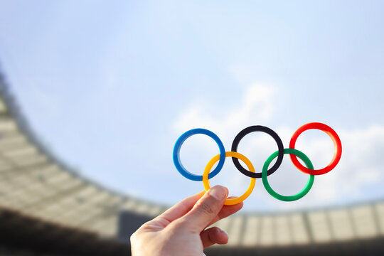 A hand holds the Olympic rings with colors representing the continents, set against the backdrop of an Olympic stadium. The image symbolizes the spirit and commencement of the Olympic Games