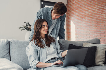 Happy millennial couple sit relax on couch in living room, smiling young husband standing from back and wife rest on sofa at home browsing Internet using modern computer device.