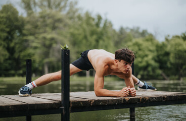 Focused young man performs a challenging exercise on a wooden dock by the water, surrounded by lush...