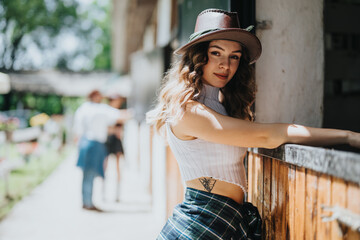 Young woman with a tattoo and wearing a hat leaning on a fence at a horse stable, capturing a...