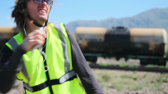 A workman wearing a helmet fast dancing while listening to music on the railway. Medium plan. Enjoy the moment as a railway worker in green uniform.