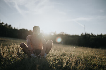 A shirtless man sits crossed-legged in a grassy field at sunset, enjoying the tranquility and...
