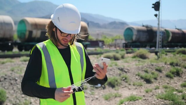 A railway inspector untangles a smartphone earpiece while standing dressed in a green reflective vest and a white helmet against the background of oil wagons.