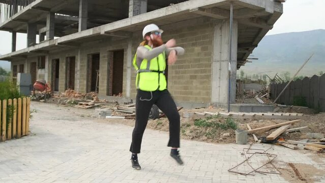 Bearded builder dancing waving his arms in sunglasses and helmet enjoying successfully completed project and the end of the working day at construction site.
