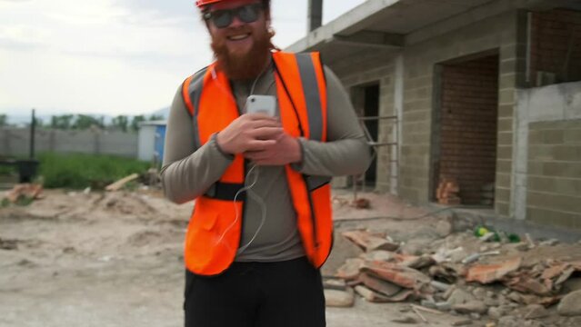 Excited male construction worker at construction site dancing enjoy moment in orange uniform and safety helmet, looking camera with smile singing to music on phone.