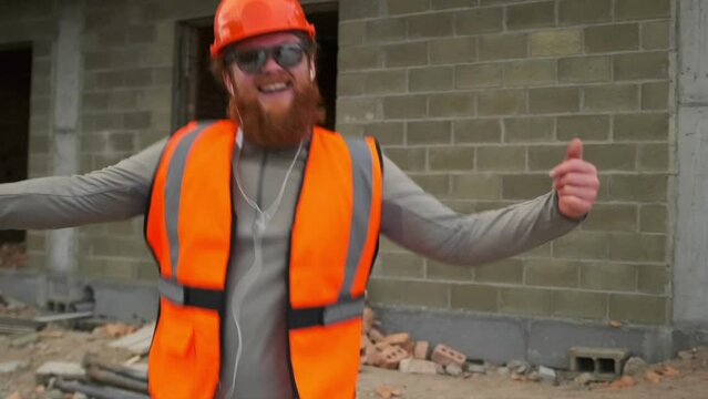 Close up dancing construction worker listen to music on headphones, construction site background, guy having fun while building house.