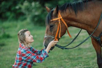 Obraz premium Happy blonde with horse in forest. Woman and a horse walking through the field during the day. Dressed in a plaid shirt and black leggings.