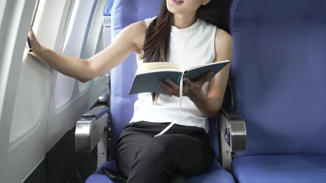 A young Asian woman, an airplane passenger, sits by the window seat, studying abroad. She gazes out the window, ready for takeoff in the economy class section.