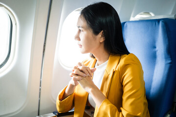 A young Asian woman, an airplane passenger, sits by the window seat, studying abroad. She gazes out...