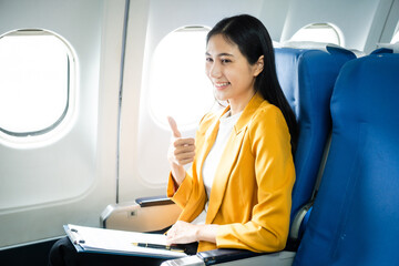 A young Asian woman, an airplane passenger, sits by the window seat, studying abroad. She gazes out the window, ready for takeoff in the economy class section.