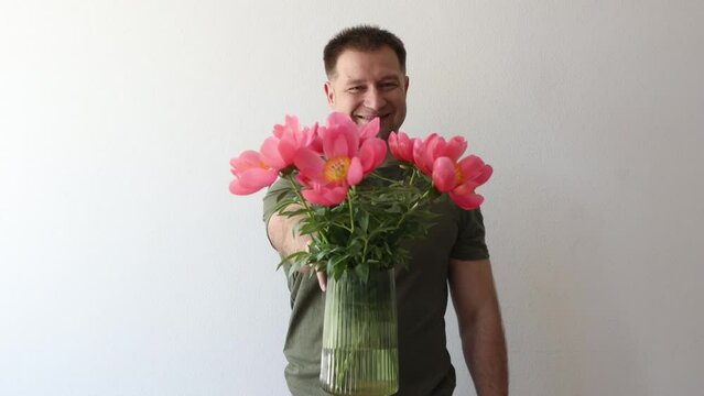 A man in a hacky t-shirt holds a vase of peonies and points to the camera