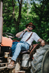 Obraz premium Relaxed farmer in a rustic outfit with a hat and suspenders sitting on a tractor while smoking cigarette in a rural setting.