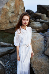 Serene woman in white dress standing on rocky shore gazes confidently at the camera by the ocean