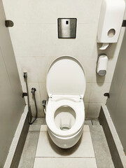 Inside a public toilet cubicle, white walls surround a toilet with a closed lid. A toilet paper...