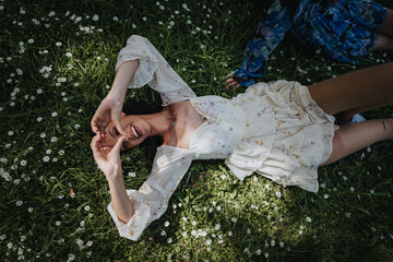 A young girl with a joyful expression lies back on the grass, surrounded by small white flowers, conveying a sense of relaxation and happiness in a park setting.