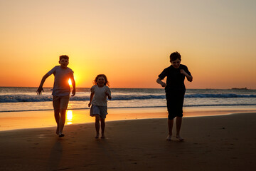 Fototapeta premium Three kids silhouettes running and jumping on beach at sunset. happy family, two school boys and one little preschool girl. Siblings having fun together. Bonding and family vacation.