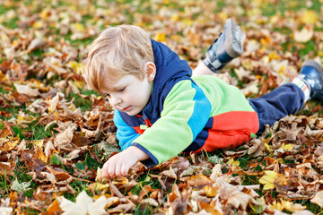 Cute little boy playing with maple leaves outdoors. Happy child walking in autumn park. Toddler baby boy wears trendy jacket . smiling Blonde boy portrait. Autumn fashion. Stylish child outside.