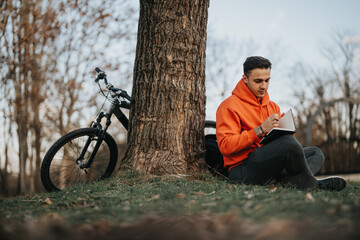 A young man with a bright orange hoodie relaxes and writes in a notebook by a tree in the park, his bike leaning nearby.